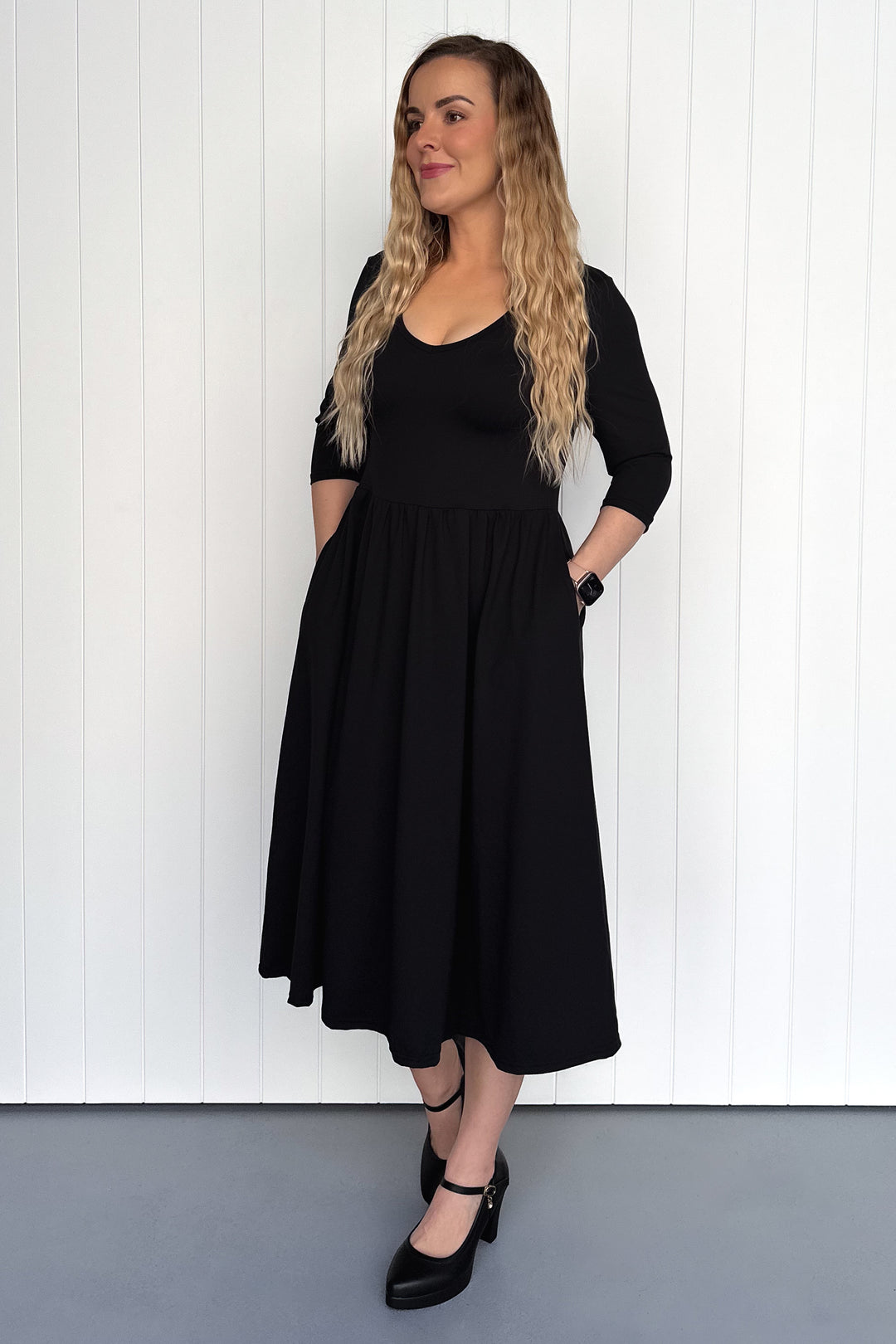 Woman wearing a black dress standing against a white paneled wall.