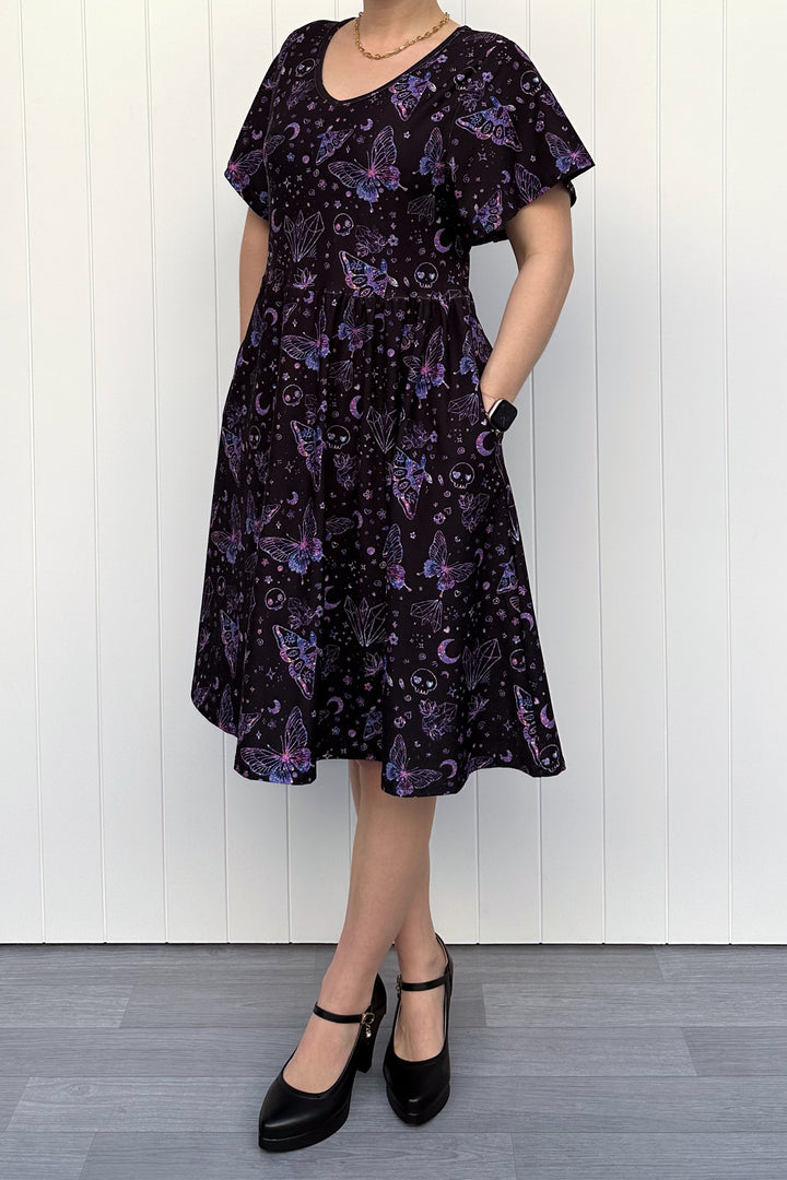 Person wearing a glitter purple dress with witch, skull and butterfly pattern against a white wooden panel background