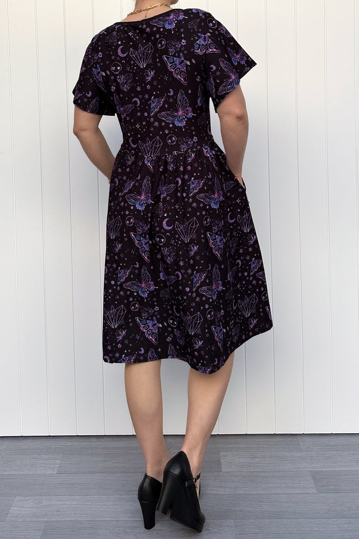 Person wearing a glitter purple dress with witch, skull and butterfly pattern against a white wooden panel background