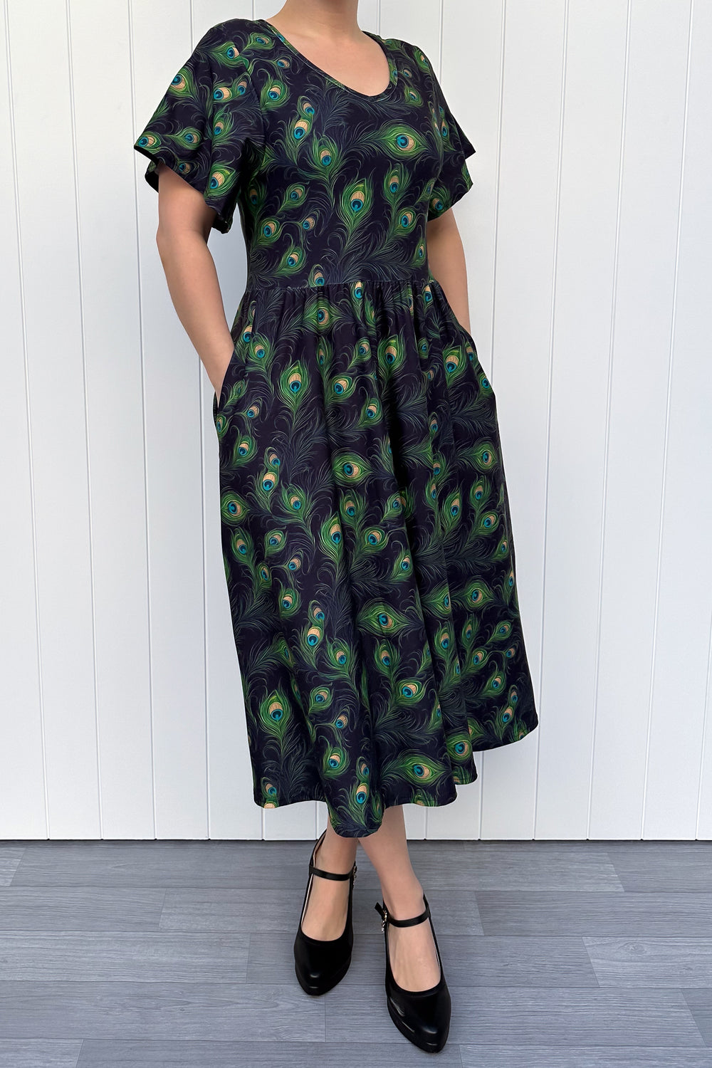 Person wearing a dark dress with peacock feather pattern against a white wooden panel background