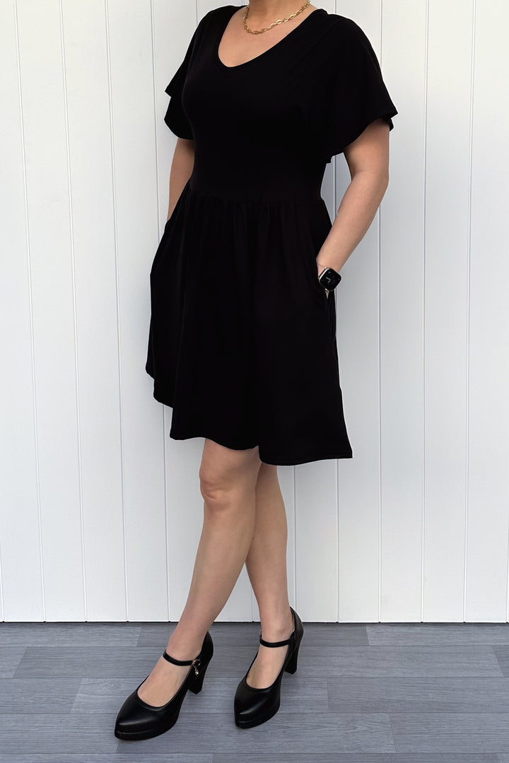 Person wearing a black dress standing against a white paneled wall.